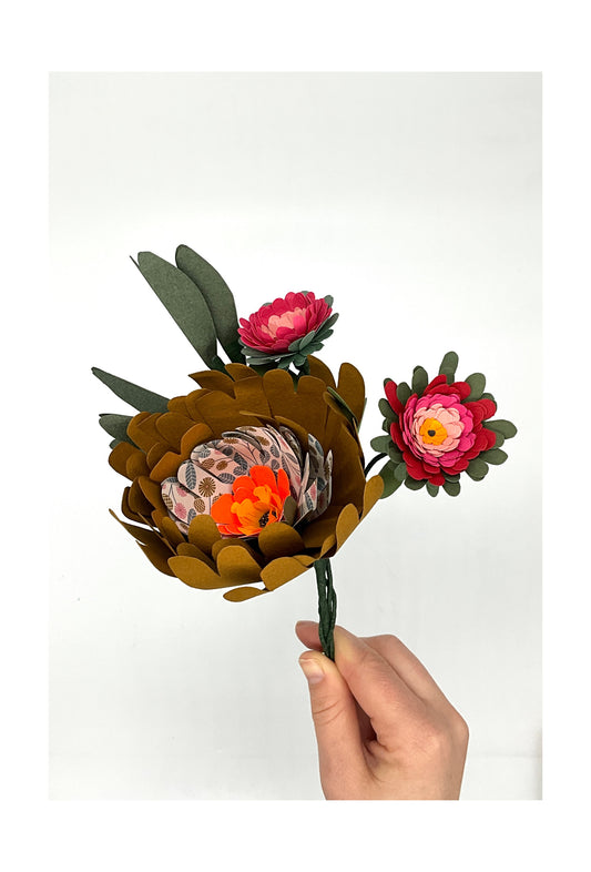Bouquet de trois fleurs en papier avec feuillage. La fleur principale est ocre