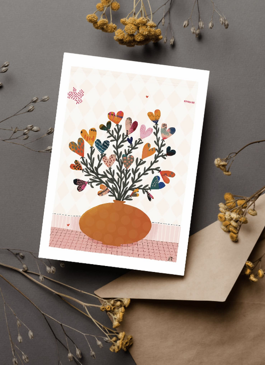 Carte postale représentant un bouquet de fleurs en forme de coeur avec de fines feuilles le long de la tige. Le bouquet est dans un vase oval de couleur rouille. Le vase est posé sur une surface quadrillé rose.